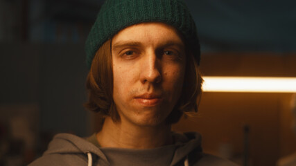 Young craftsman in hoodie and hat looking at camera while standing in dimly illuminated sculpting studio