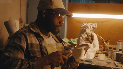 African American craftsman in checkered shirt and cap polishing half of plaster bust while sitting near illuminated workbench and working in sculpting workshop at night