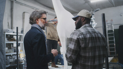 Low angle of mature customer in suit discussing gypsum giraffe statue with black craftsman in illuminated workroom