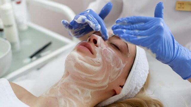 Beautician doctor is applying facial cleansing foam on woman's face. Facial skin massaging in cosmetology clinic