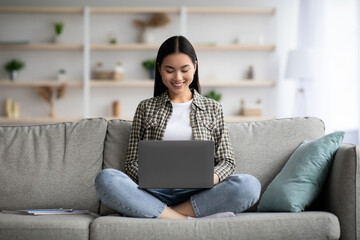 Smiling asian woman typing on laptop keyboard, home interior