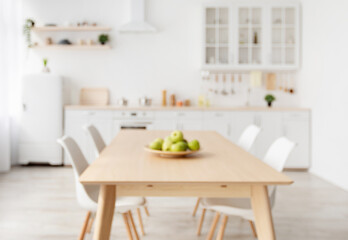 Blurred background with modern light kitchen, wooden dining table and white furniture, chairs, kitchenware and utensils