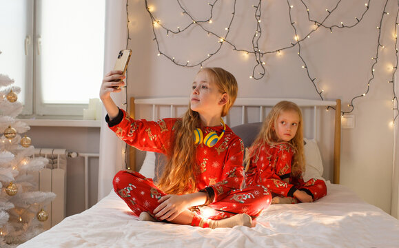 beautiful little girls are sitting in bed in bright Christmas pajamas taking pictures of themselves on the phone, a bright room. Christmas. New Year