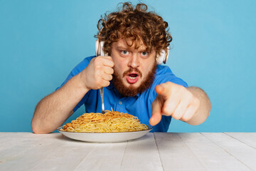 Close-up portrait of young hairy red-bearded man sitting at table with large portion of noodles, pasta isolated on blue studio background.