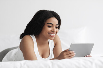 Watching video and news. Excited black woman holding digital tablet and lying on bed in the morning, free space