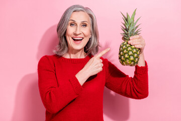 Portrait of attractive trendy cheerful gray-haired woman demonstrating ananas isolated over pink...