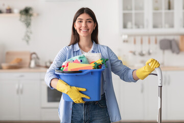 Cheerful young housewife holding bucket with cleaning supplies