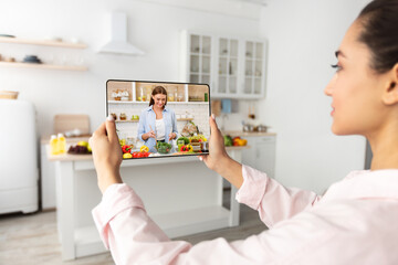 Young lady watching online culinary tutorial at kitchen, using tablet pc, studying remotely during...