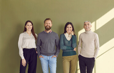 Group portrait of four happy people in smart casual outfits posing against green studio background. Team of 4 confident business men and women standing by office wall, smiling and looking at camera