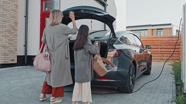 Tracking medium shot of young mother and 9-year-old daughter loading their bags into trunk of electric car charging before house in suburbs