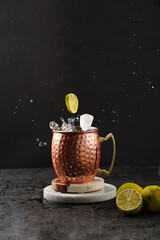 Traditional american alcoholic beverage moscow mule in copper mug with flying lemon and ice cube on white marble boards, black background