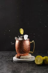 Plexiglas foto achterwand Traditional american alcoholic beverage moscow mule in copper mug with flying lemon and ice cube on white marble boards, black background © Romana