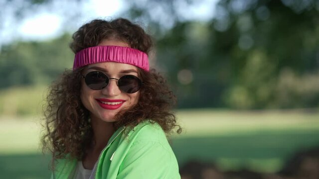 Close-up face smiling Caucasian retro woman putting on sunglasses looking at camera. Headshot portrait cheerful confident attractive lady in hair band posing outdoors in sunny summer park