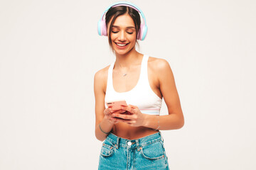 Beautiful smiling woman dressed in jeans clothes. Sexy carefree model listening music in wireless headphones. Adorable and positive female posing on grey background in studio. Holding smartphone