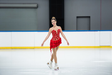 full length of cheerful woman in red dress figure skating on professional ice rink