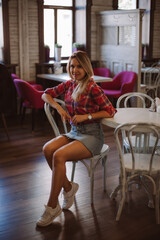 Obraz na płótnie Canvas Adult shining blonde woman with face looking into camera on chair in cafe leaning on chair back with left hand wearing checkered shirt tied in belly area, jeans skirt and white sneakers in daytime.
