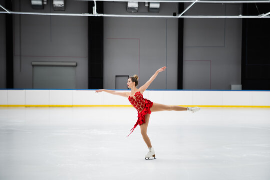 full length of figure skater in red dress performing camel spin in professional ice arena