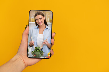 Female hand holding cellphone with online culinary class on screen over orange background, panorama with copy space