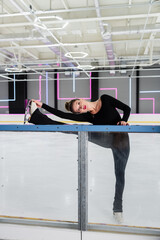 full length of professional figure skater in black bodysuit and ice skates stretching near frozen...