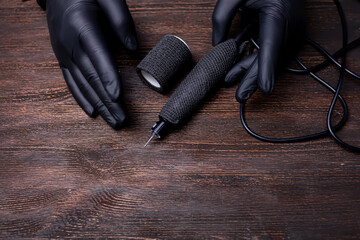 the hands of a permanent makeup artist in black sterile gloves in front of which lies a tattoo machine wrapped with a bandage tape on a wooden table - obrazy, fototapety, plakaty