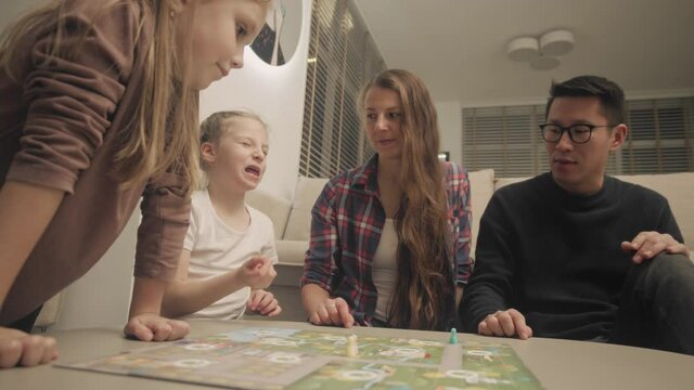 Diverse Family With Two Little Girls Playing Board Game In Living Room In The Evening. One Girl Is Shaking Dice And Make Her Move With Facial Expression