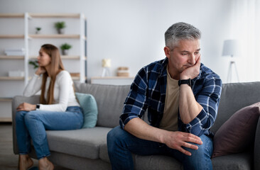 Offended mature man and woman sitting on different sides of sofa after after quarrel indoors, empty space - obrazy, fototapety, plakaty