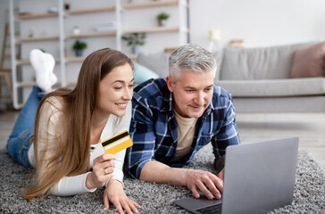 Cheerful mature spouses ordering goods online with laptop pc and credit card, lying on floor at home