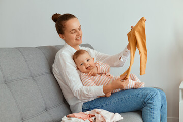 Portrait of smiling young adult mother sitting on sofa with her toddler daughter, holding baby's clothing bought in store, spending time with her child, expressing positive emotions.