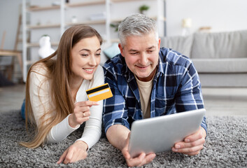 Cheerful mature couple lying on floor with tablet computer, holding credit card, making payment together in online store