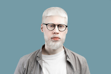 Portrait of serious albino man with pale skin and blue eyes, wearing eyeglasses against turquoise...