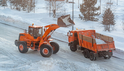 Big orange tractor cleans up snow from the road and loads it into the truck. Cleaning and cleaning...