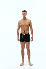 Full-length portrait of young handsome shirtless sportive man wearing black boxer-briefs standing isolated on white studio background.