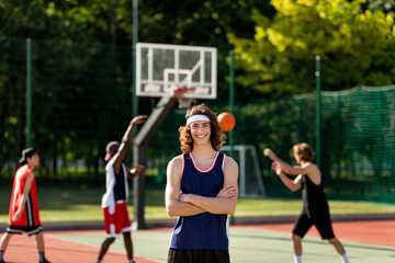 Happy Caucasian sportsman posing with crossed arms and smiling at camera, his diverse team playing basketball, outdoors