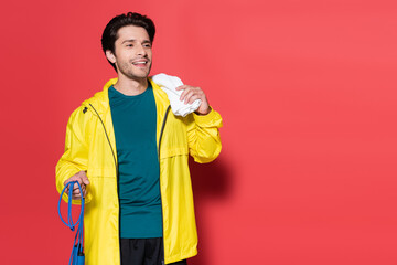 Smiling sportsman holding jump rope and towel on red background