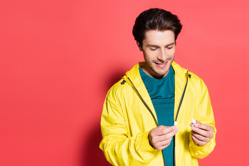 Cheerful sportsman holding wireless earphones on red background