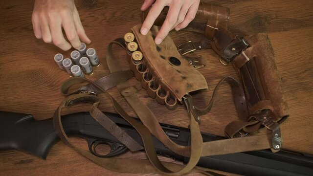 man is  preparing for hunting and fills a bandolier with a 12 gauge cartridges near a black shotgun on the table
