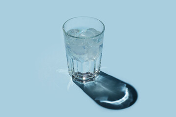 Transparent glass of water with ice cubes and shadow on blue background.