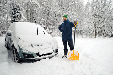 Man with shovel standing near snow covered car after heavy snowfall. Bad winter weather, natural...