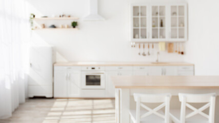 Sunny kitchen interior in Scandinavian style with white kitchen furniture and dining area, blurred background