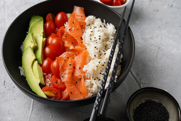 Poke bowl - black bowl with white rice, smoked salmon, cherry tomatoes and avocado, sesame seeds, soy sauce and chopsticks on a grey concrete surface