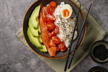 Poke bowl - green bowl with white rice, smoked salmon, cherry tomatoes and avocado, sesame seeds, soy sauce and chopsticks on a grey concrete surface, top view