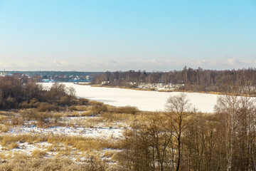 Panoramic winter landscape view with frozen river or lake covered with ice, reeds and some trees off the coast, small village and tower in background