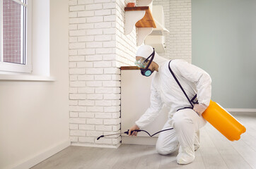 Pest control guy inside the house. Male in white protective overalls crouching by kitchen wall and...