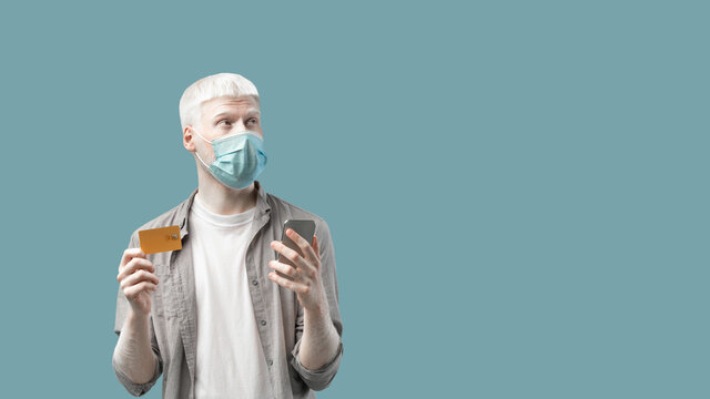 Extraordinary man in face mask holding smartphone and credit card, looking at empty space on turquoise studio background - Powered by Adobe