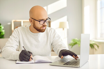 Serious man in glasses and comfortable fingerless compression hand gloves for easing arthritis pain sitting at desk, using computer, typing on keyboard, holding pen and taking study notes on paper - obrazy, fototapety, plakaty