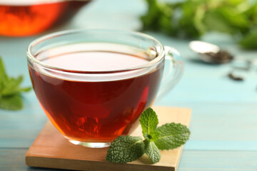 Glass cup of aromatic black tea with fresh mint on light blue wooden table against blurred background, closeup