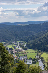 Obraz premium Aerial view, town of Bourscheid with its houses between a country road and the river, countryside with mountains with lush green trees in the background, summer day with a cloudy sky, Luxembourg