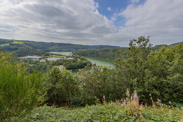Fototapeta na wymiar Valley with the Esch-sur-Sure lake and the dam surrounded by wild vegetation and lush green trees seen from a viewpoint, sunny day with a blue sky and abundant white clouds in Luxembourg