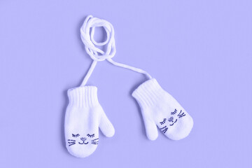 Very Peri Color of the year 2022. Violet. White knitted baby mittens isolated on a purple background, top view. Winter kids clothes. Mittens with Smile. Little baby gloves