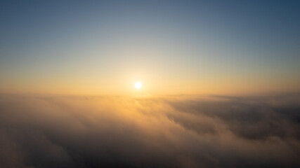 Aerial View. Flying in fog, fly in mist over the early morning clouds in the rising sun. Aerial camera shot. Flight above the clouds towards the sun with the mist clouds floating by. Misty weather